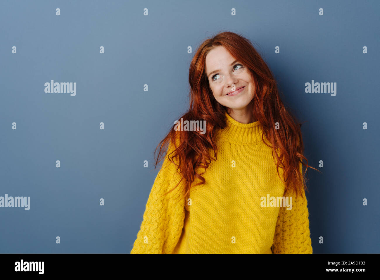 Simpering giovane donna con un sorriso coy cercando da parte con un sorriso come lei flirta su un blu di sfondo per studio con spazio di copia Foto Stock