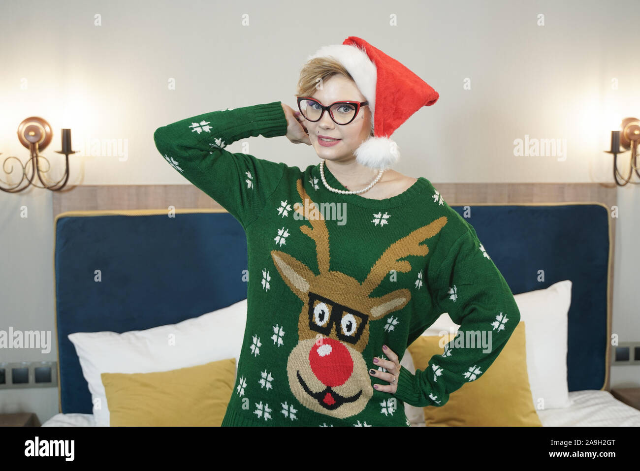 Carino dimensione plus nerd ragazza in bicchieri e verde maglione di Natale con le renne e rosso di Santa Claus hat al mattino sul letto in posa. Foto Stock