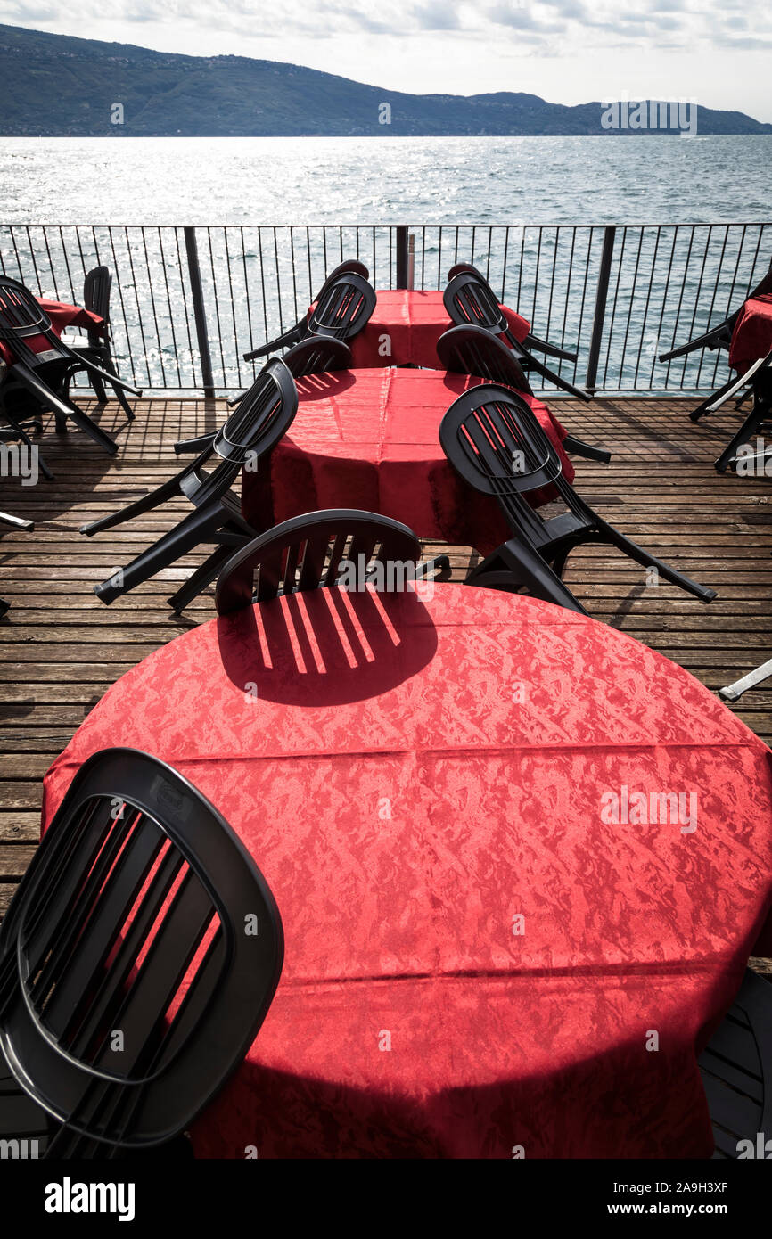 Sedie nere sono ribaltati contro l'esterno cafe tabelle con colore rosso brillante tovaglie sui decking accanto al Lago di Garda Foto Stock