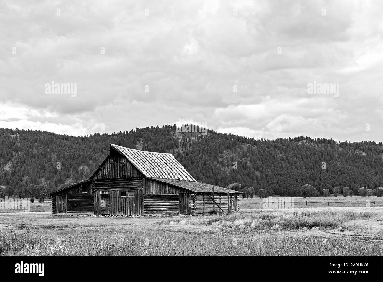 Vecchio fienile pioniere in campo vuoto con montagne oltre sotto cielo nuvoloso, nero e bianco. Foto Stock