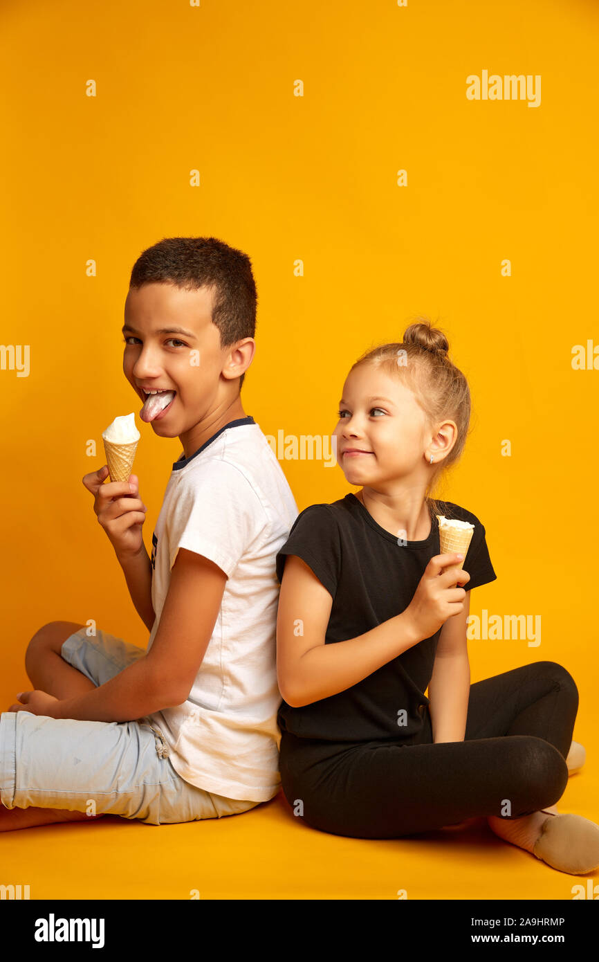 Divertente i bambini mangiano gelato alla vaniglia in un cono di cialda su uno sfondo giallo, gioiosa del fratello e sorella Foto Stock