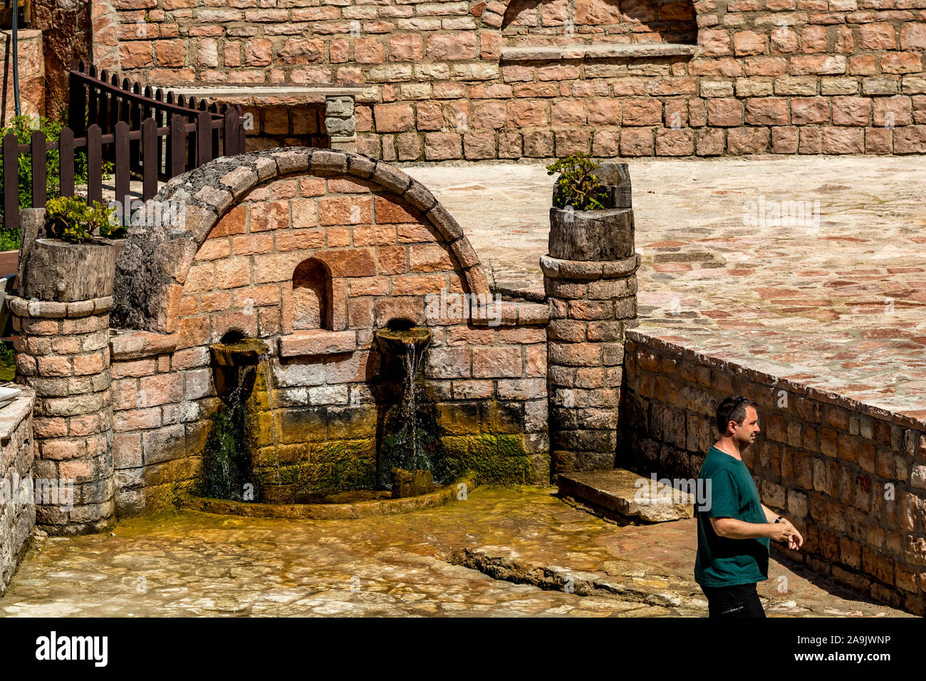 KRONGJI, ALBANIA - Giugno 7, 2019: Maschio turistico con maglietta verde passando da muro di pietra rubinetto di acqua ed esecuzione di bere acqua limpida, Blue Eye nazionale parco naturale all'aperto, Albania, soleggiata giornata di primavera Foto Stock