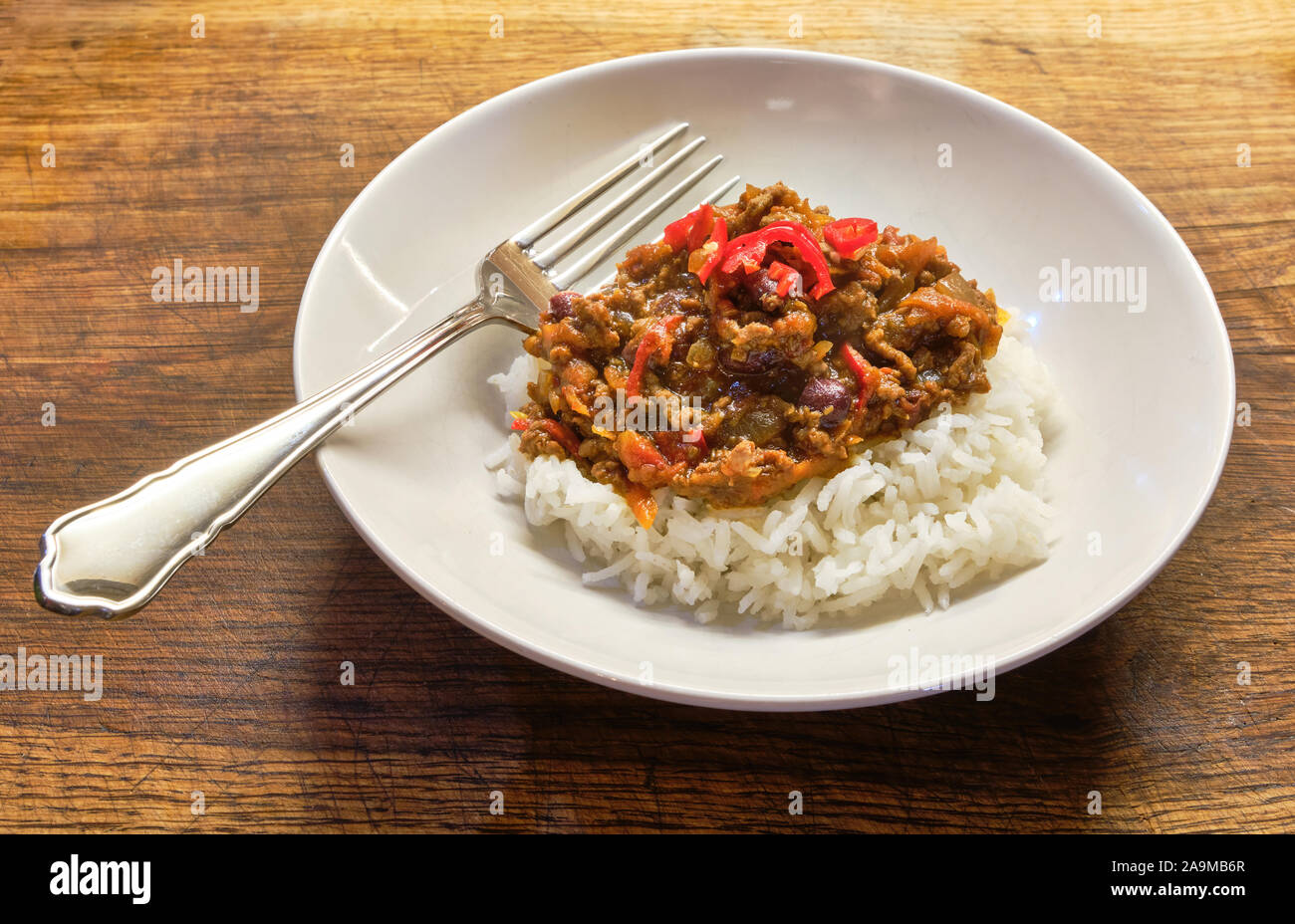 Una ciotola bianco della casa di fresco cucinato chili con carne e riso con una forcella guarnita con rosso peperoncino tritato pepe e posto su una tavola di legno Foto Stock