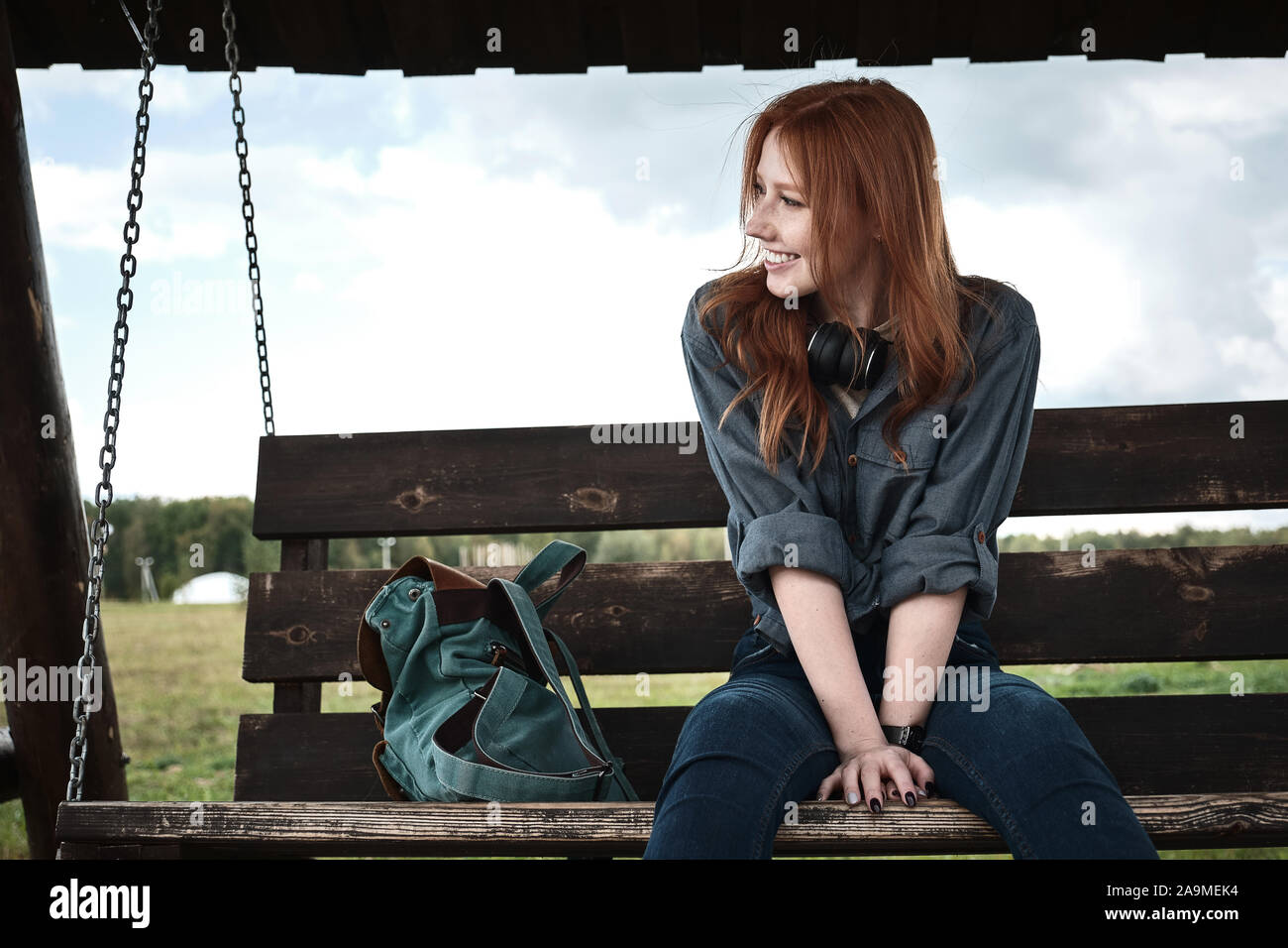 Redhead girl in un paio di jeans shirt si siede con uno zaino su una oscillazione di legno da banco, sporgersi in avanti e sorridente, guardando al lato. Foto Stock