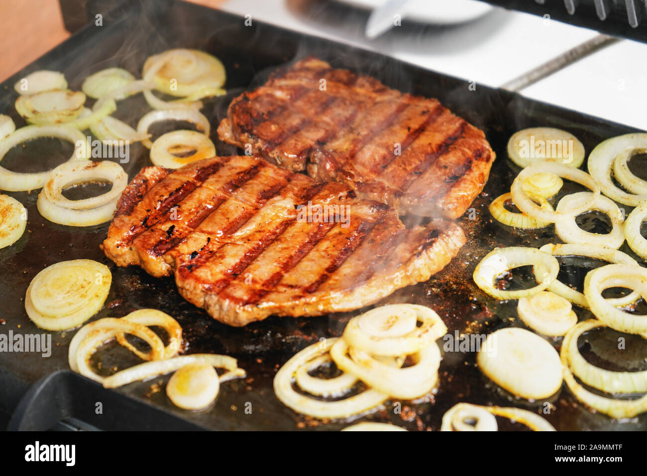 Due costolette di maiale bistecche e la cipolla tagliata a fette e grigliate sulla griglia elettrica, fumo di cibo alla griglia visibile Foto Stock