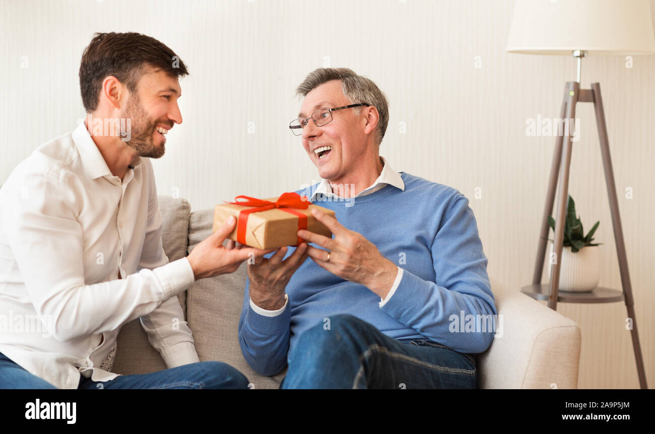 Coppia figlio congratularmi con anziani padre dando dono seduto sul divano Foto Stock