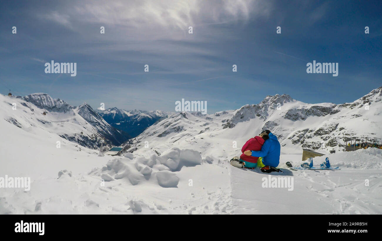 Un paio di sci abiti, seduto sulla neve, avvolgente e godendo le montagne innevate. Infinite gamme di neve montagne tappata davanti a loro. A Foto Stock
