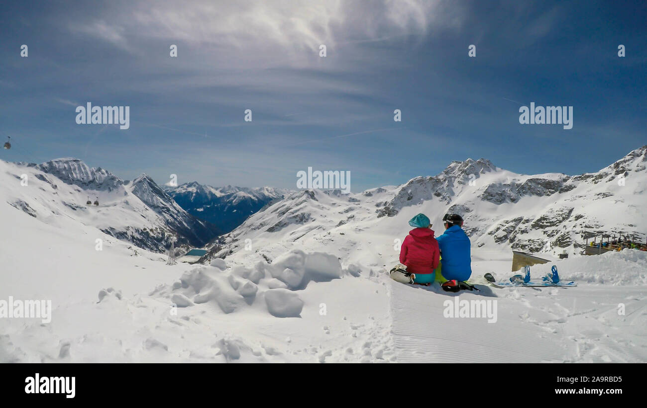 Un paio di sci abiti, seduto sulla neve, avvolgente e godendo le montagne innevate. Infinite gamme di neve montagne tappata davanti a loro. A Foto Stock