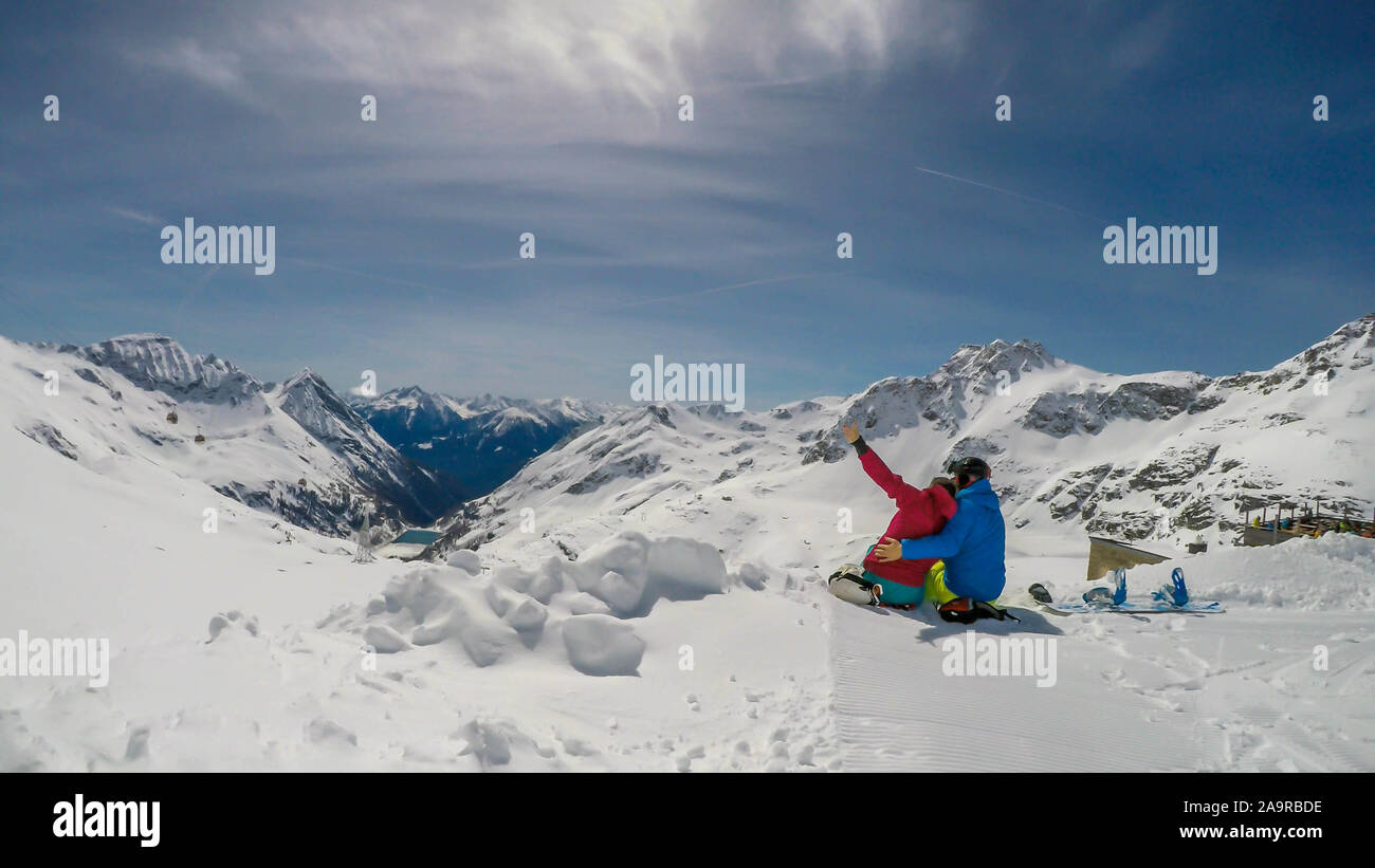 Un paio di sci abiti, seduto sulla neve, avvolgente e godendo le montagne innevate. Infinite gamme di neve montagne tappata davanti a loro. A Foto Stock