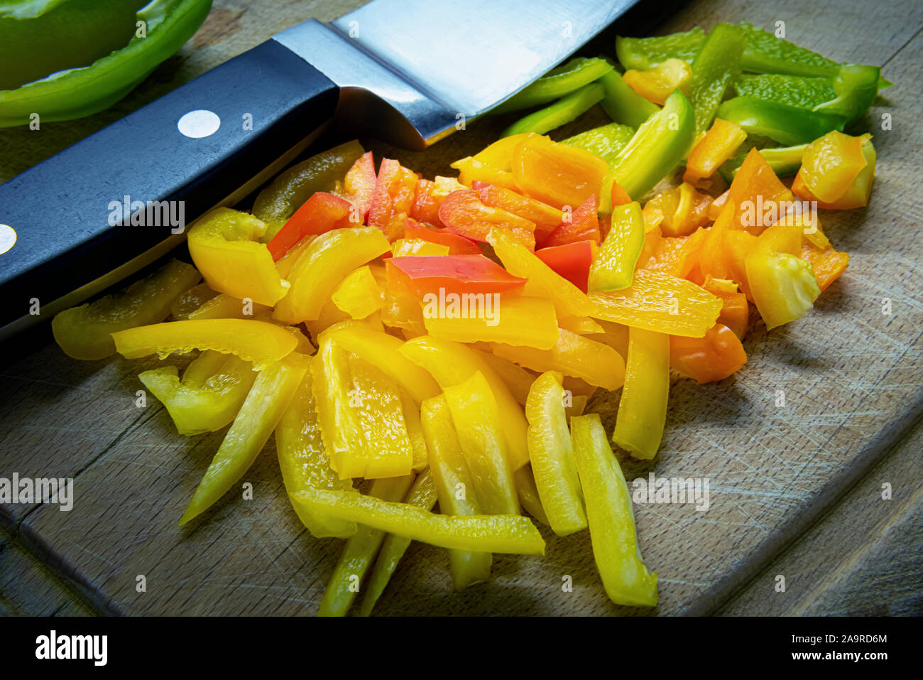 Affettato colorato di rosso, giallo, verde e arancione o campana di peperone dolce sul vecchio usato graffiato tagliere di legno. Grande coltello di metallo è vicino a pezzi di pep Foto Stock