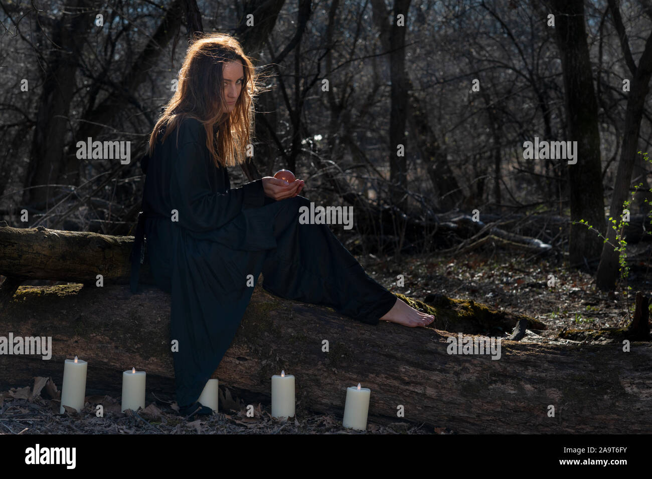 Donna con scuri capelli lunghi in black robes circondato da white candele accese seduto su un tronco di albero nella foresta. Torna al concetto di natura. Stregoneria Foto Stock