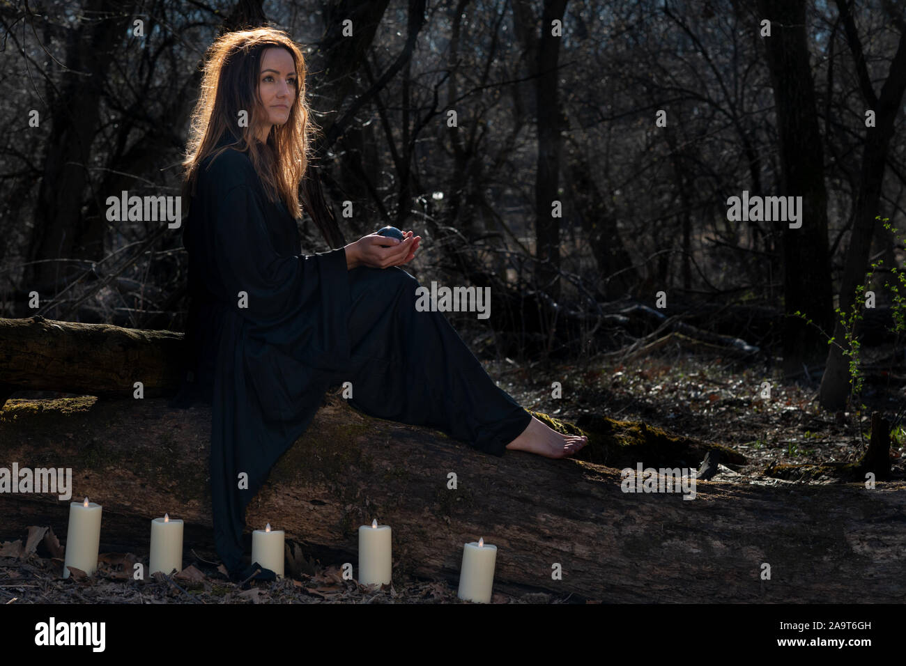 Donna con scuri capelli lunghi in black robes circondato da white candele accese seduto su un tronco di albero nella foresta. Torna al concetto di natura. Stregoneria Foto Stock