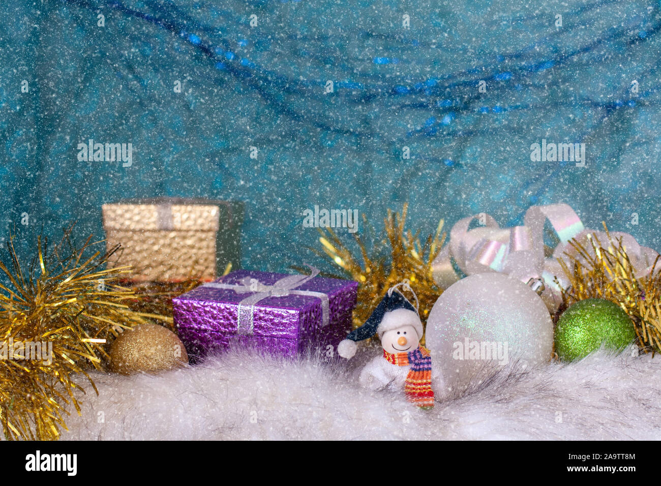 Un sorridente pupazzo di neve su una pelliccia bianca base accanto a palle di Natale e confezioni regalo. Sfondo blu con l'effetto di caduta di neve. Messa a fuoco selettiva. Foto Stock