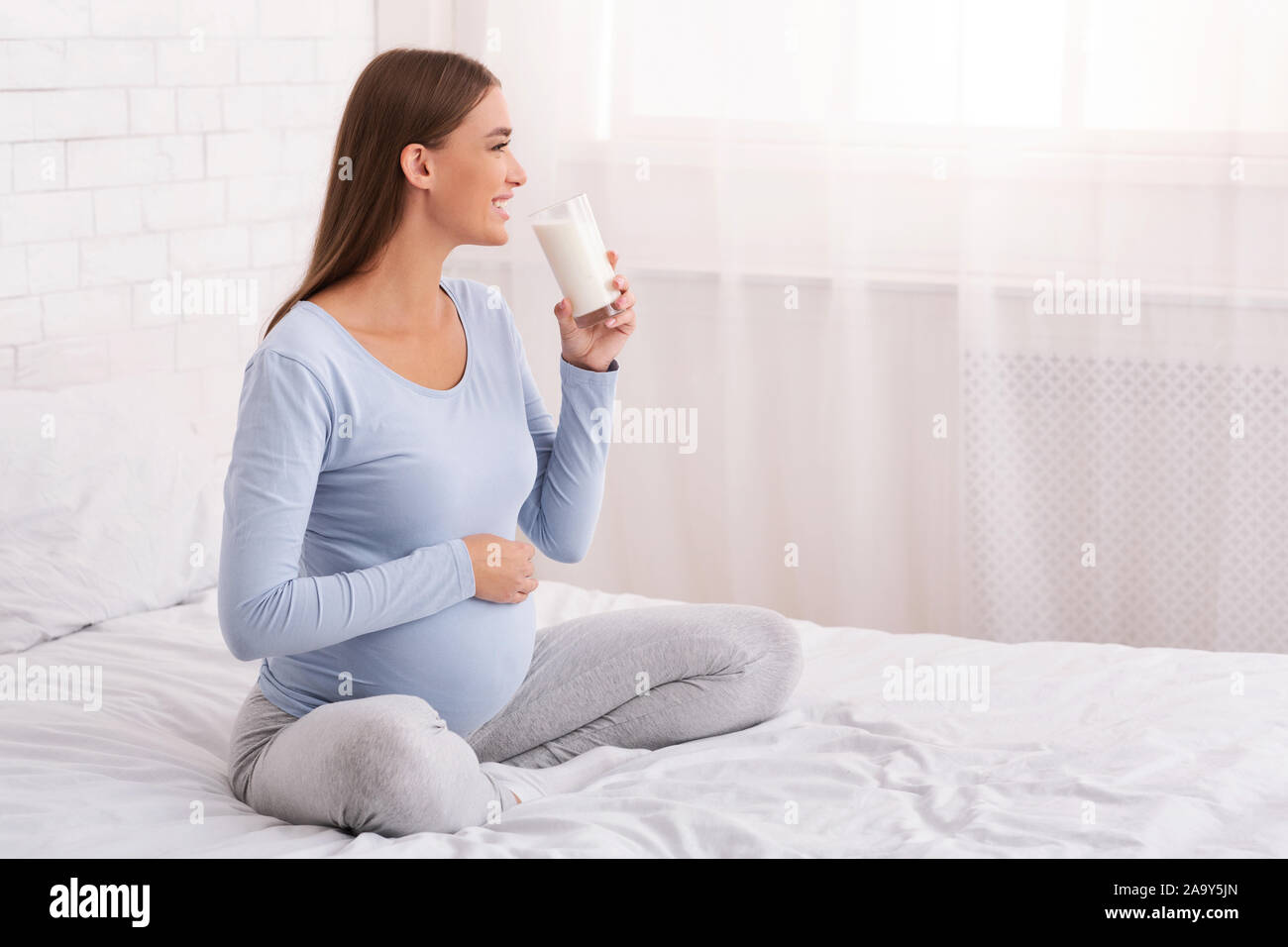 Donna incinta bere latte seduta sul letto di casa Foto Stock