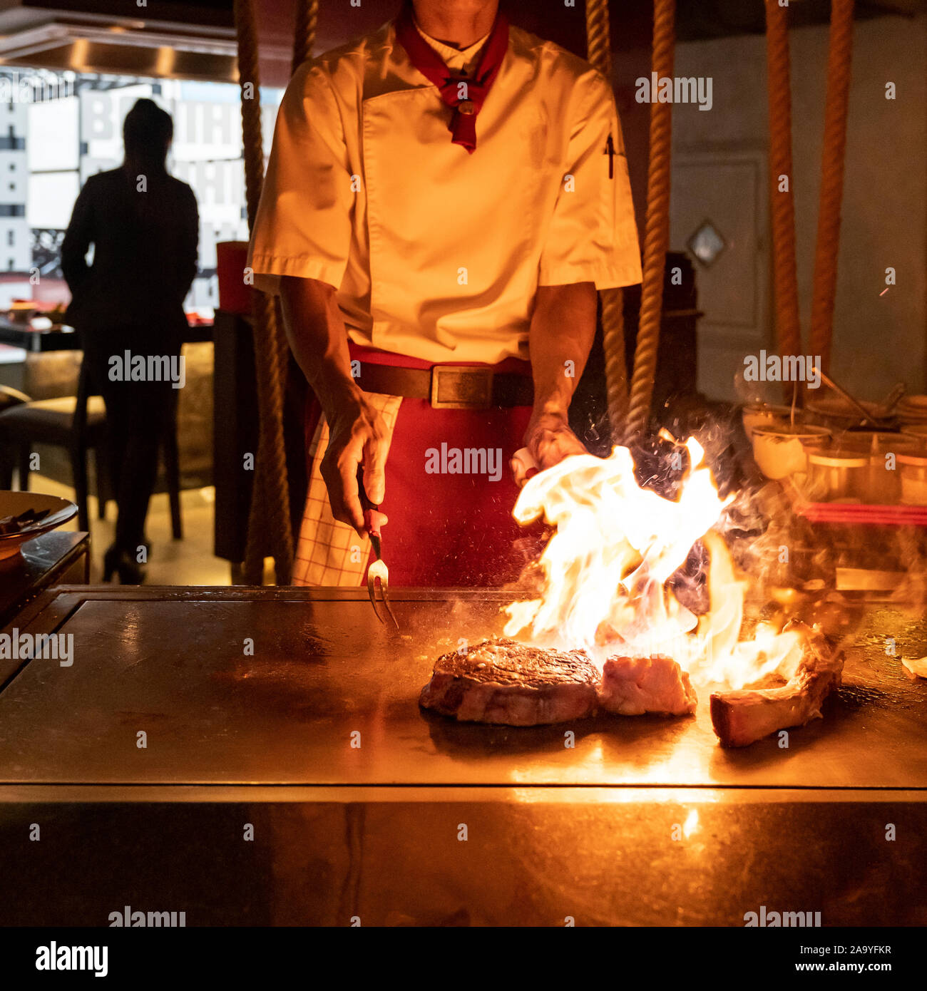 La cottura della carne di manzo ricetta teppanyaki Giapponese grigliate di carni bovine tomahawk bistecca nel recipiente di cottura utilizzando il raschiatore con fiamma in background Foto Stock