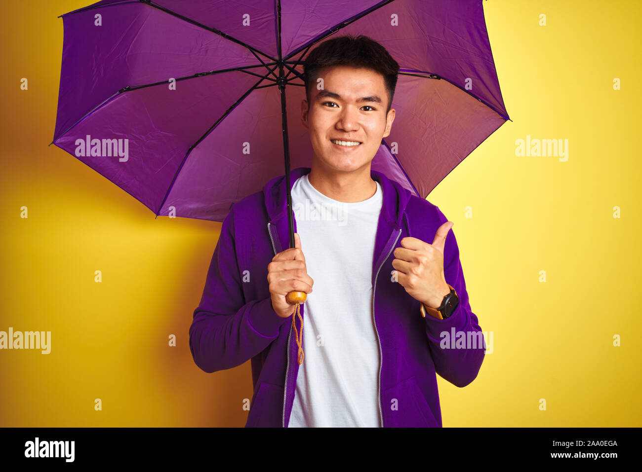 Asian uomo cinese indossando ombrello viola in piedi isolato su sfondo giallo felice con un grande sorriso facendo segno ok, pollice in alto con le dita, excellen Foto Stock