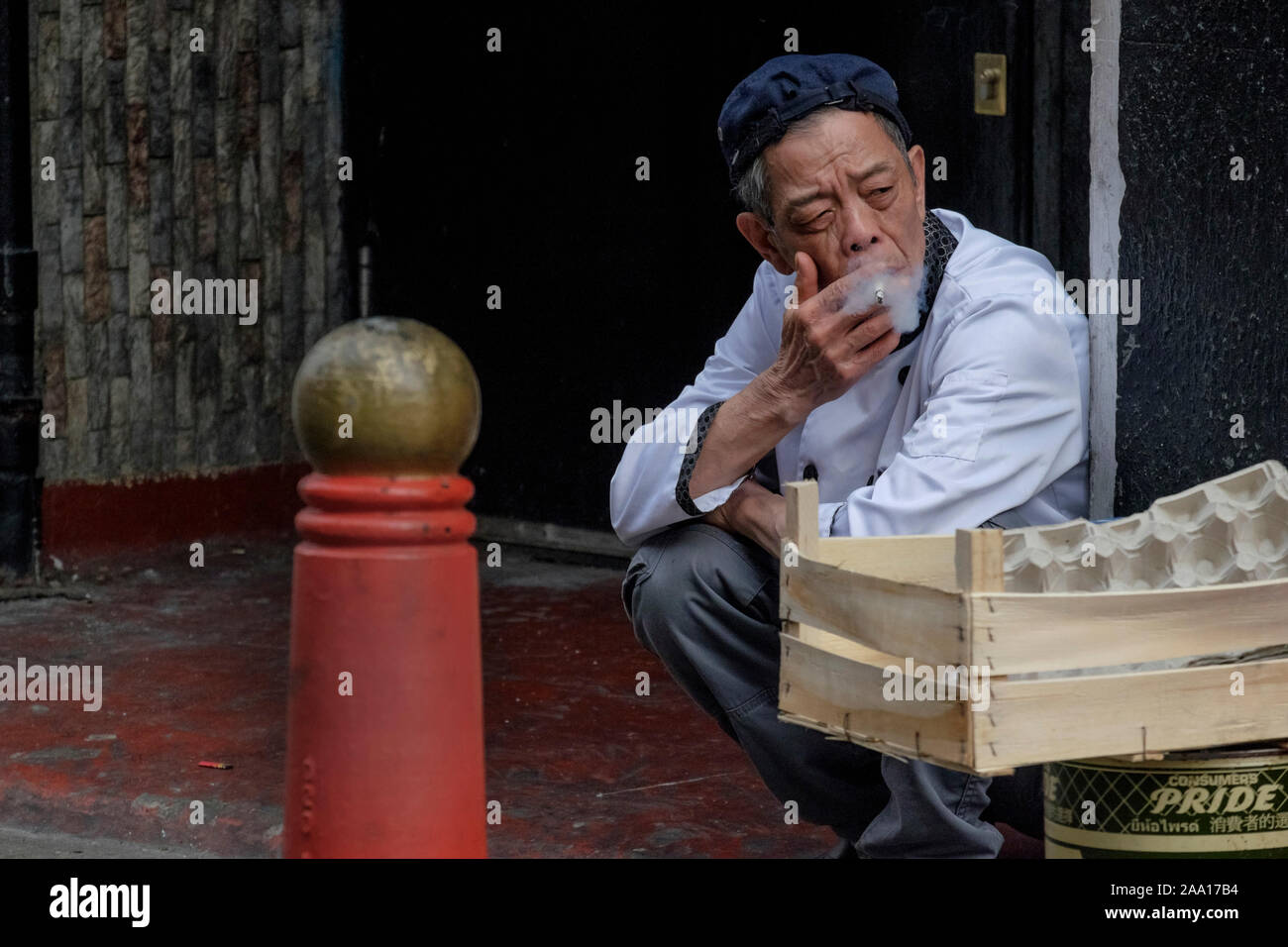 London street photography: Anziani ristorante cinese lavoratore accovacciato tenendo pausa sigaretta, Chinatown, Londra. Foto Stock