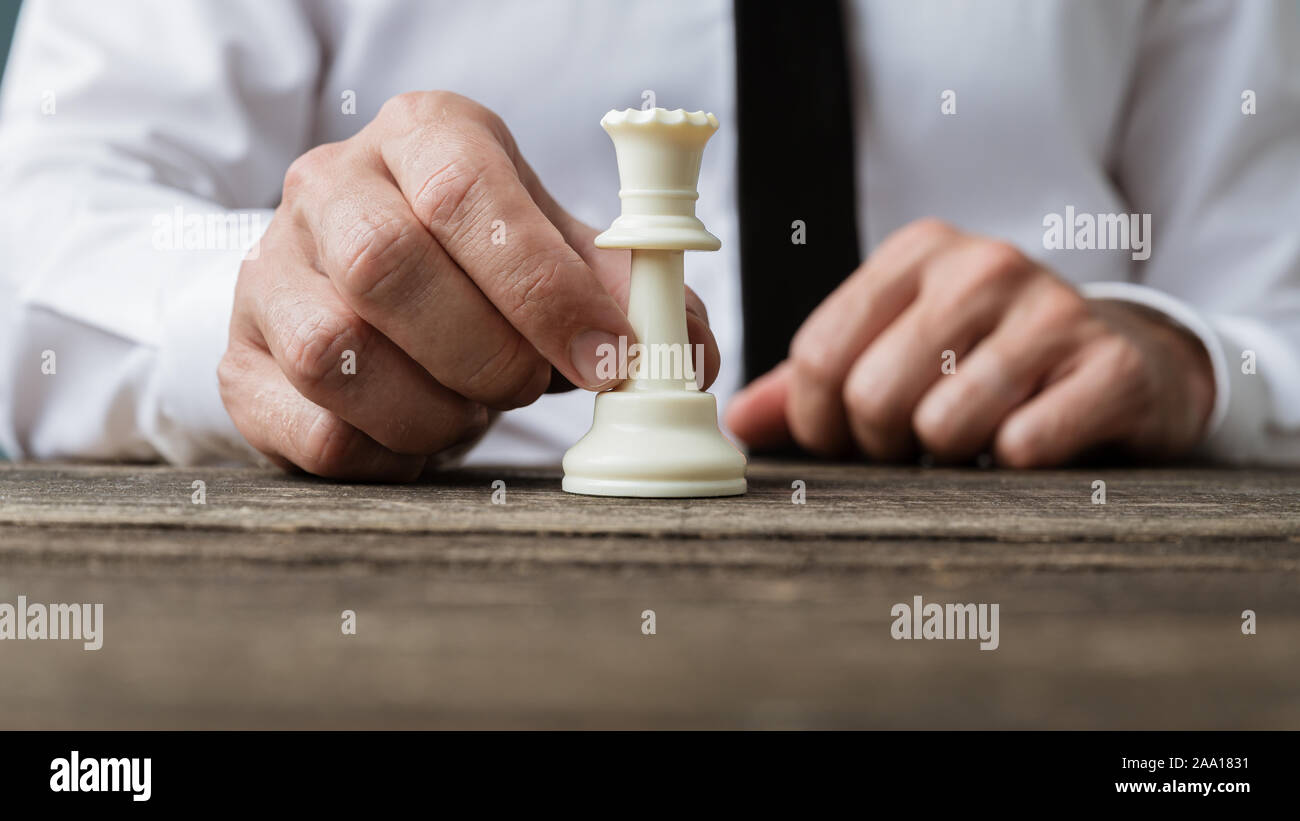 Primo piano di imprenditore tenendo un bianco a scacchi la figura del re davanti a lui per una rustica scrivania in legno in una immagine concettuale. Foto Stock