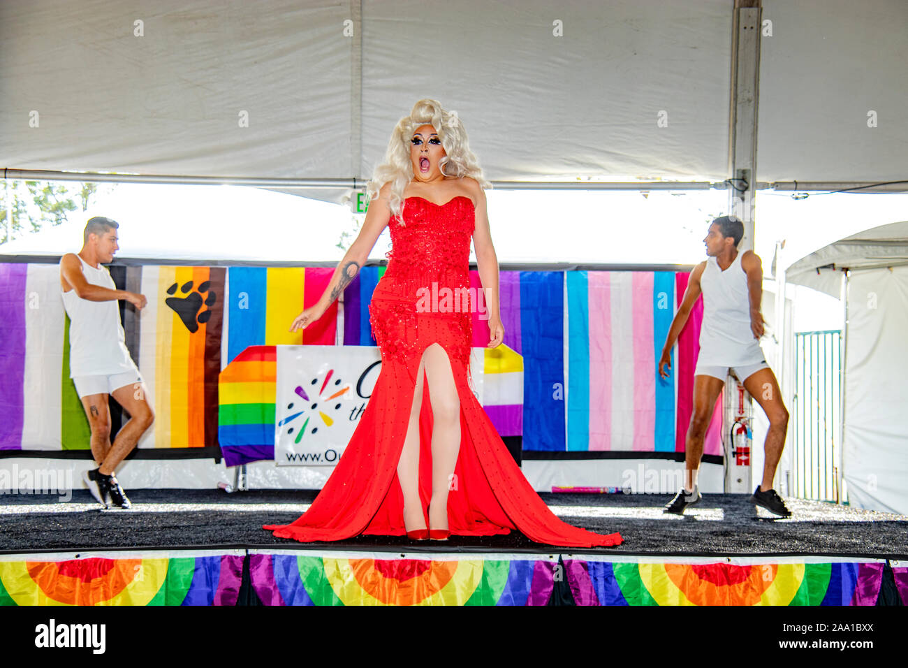 Indossa un rosso abito sequined, una drag queen suona su un palco di un County Fair LBGTQ evento in Costa Mesa, CA, fiancheggiata da due ballerini di sesso maschile. Foto Stock