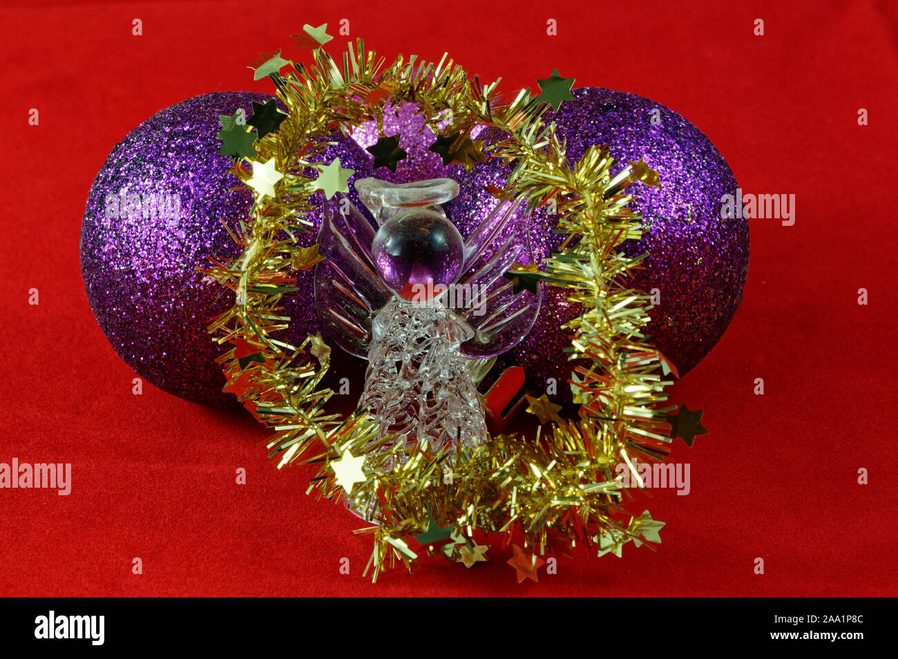 Un Natale di vetro angel è circondato da gold tinsel e si trova di fronte il viola baubles su uno sfondo rosso Foto Stock