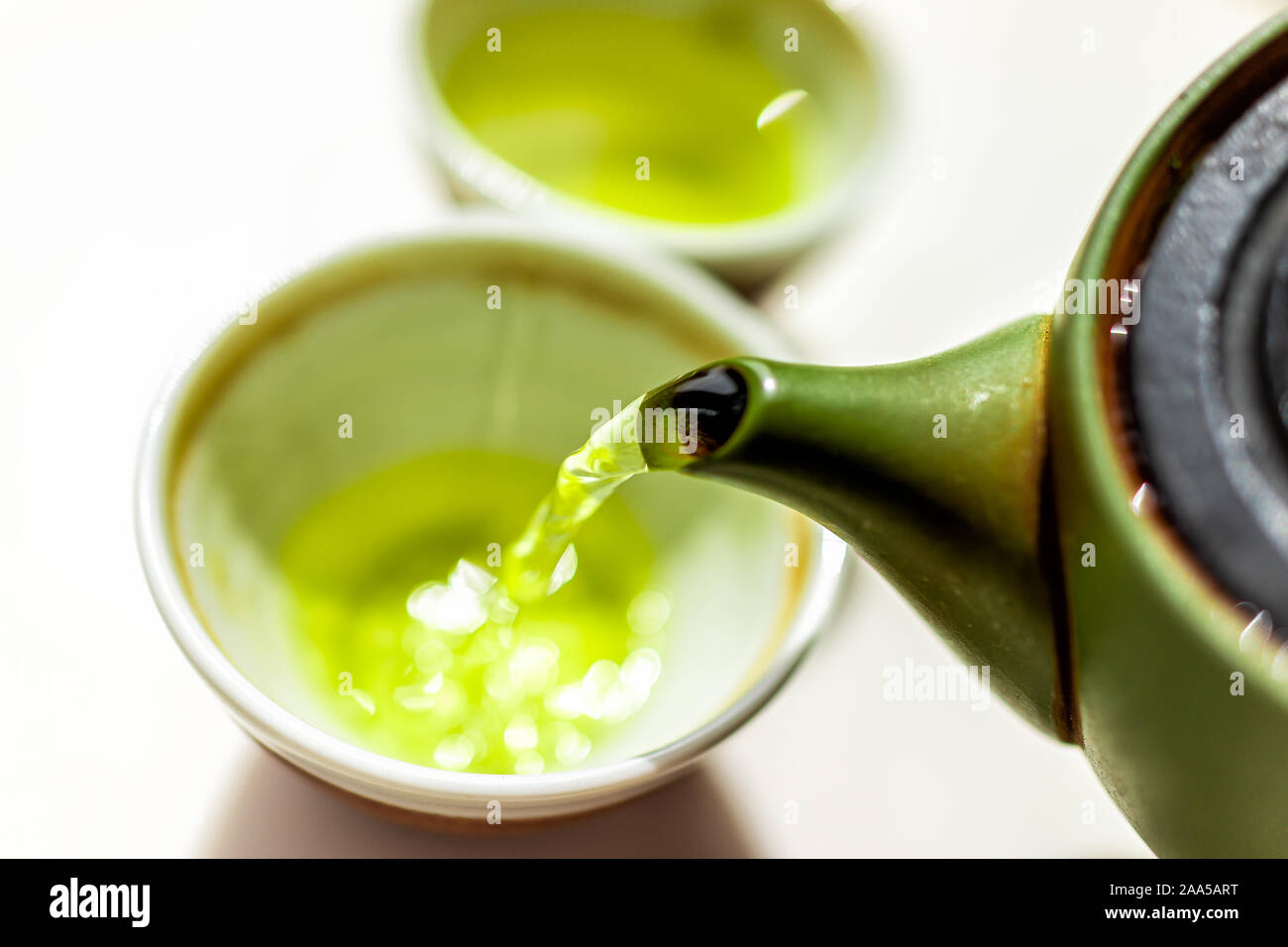 Primo piano di argilla verde teiera teiera in bianco su sfondo tabella e versando il moto del liquido colorato e vivace sencha giapponese o genmaicha drink durante Foto Stock