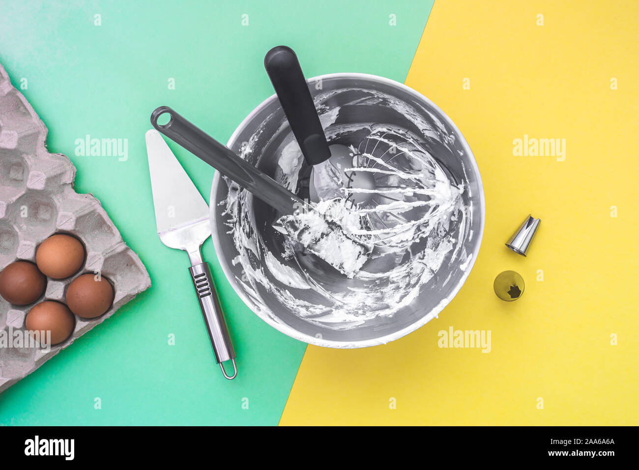 Cottura per torte utensili da effettuare nel recipiente di miscelazione dopo la torta facendo, ugelli, uova crude sul piatto verde laici sfondo giallo Foto Stock