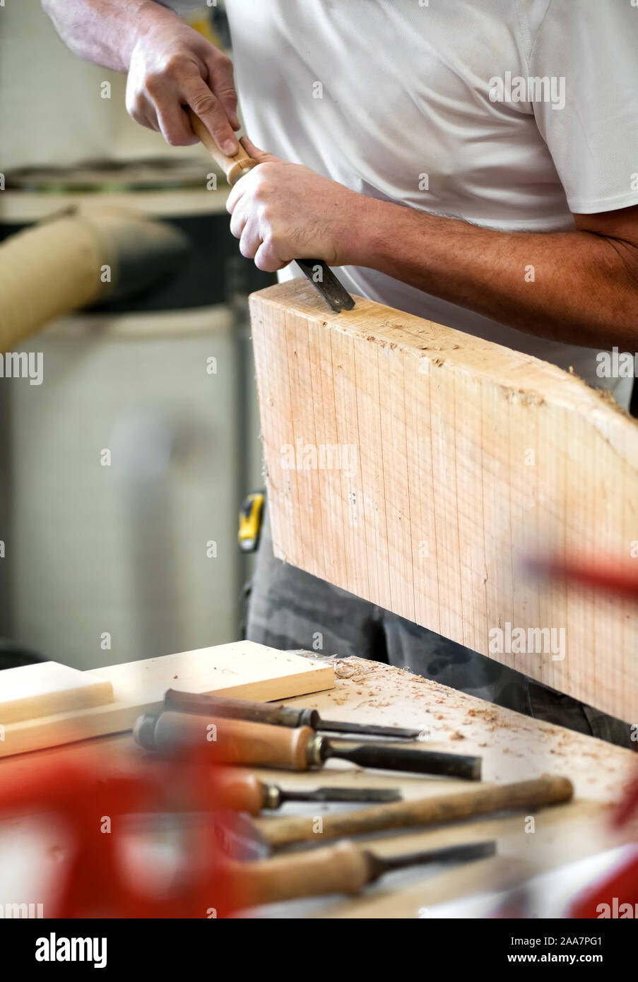 Carpenter lavorando su un blocco di legno con uno scalpello in stretta fino alle mani con un assortimento di utensili a mano sul banco in primo piano Foto Stock