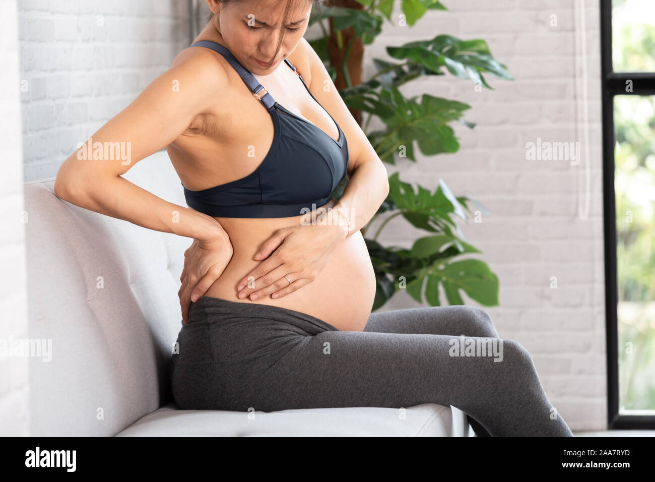 Vista laterale di sconvolgere Asian donna incinta toccando dietro e di lato che soffrono di mal di schiena e dolore anca in appartamento di luce Foto Stock
