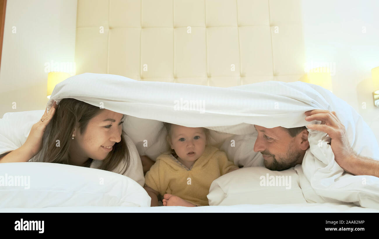 Una bella famiglia multietnica giace sul letto e si nasconde sotto una coperta. Si stanno divertendo e scherzando. Foto Stock