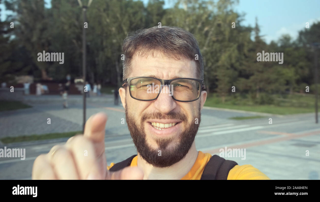 Felice di blogger maschio con gli occhiali e con una barba parla alla fotocamera sulla strada. Intorno alla gente camminare sul lungomare Foto Stock