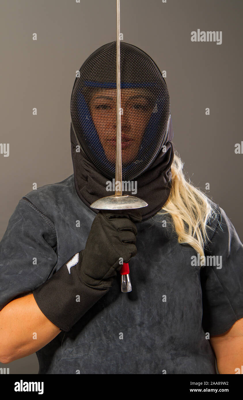 Giovane ragazza bionda in un recinto trainer uniforme scuro e nero maschera casco stand con una piattina di pinza su sfondo grigio Foto Stock