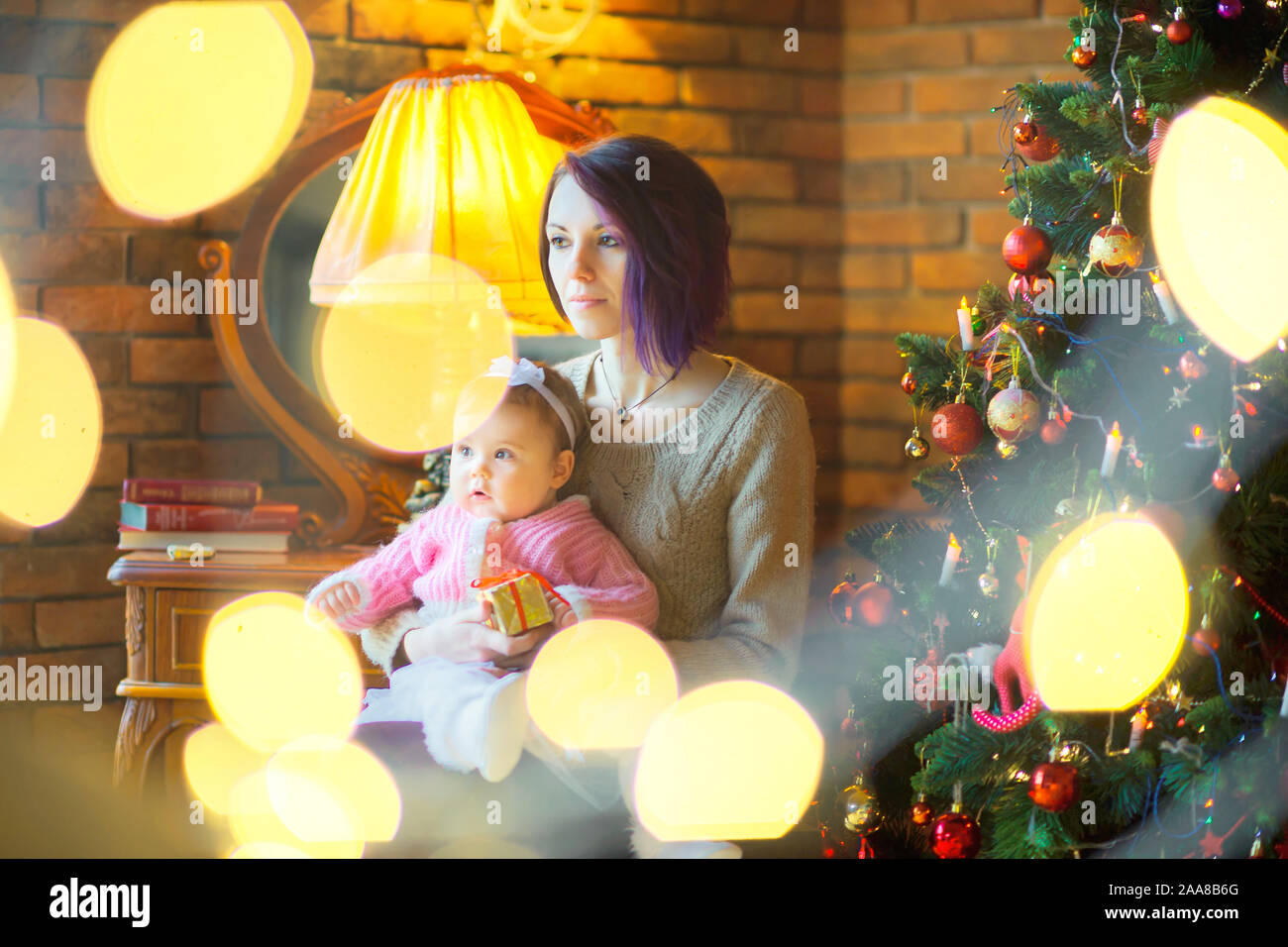 La mamma con una piccola figlia si siede vicino a una festa di Natale albero su di uno sfondo di luci luminose, tenere un piccolo dono e sogno. Vacanze Foto Stock