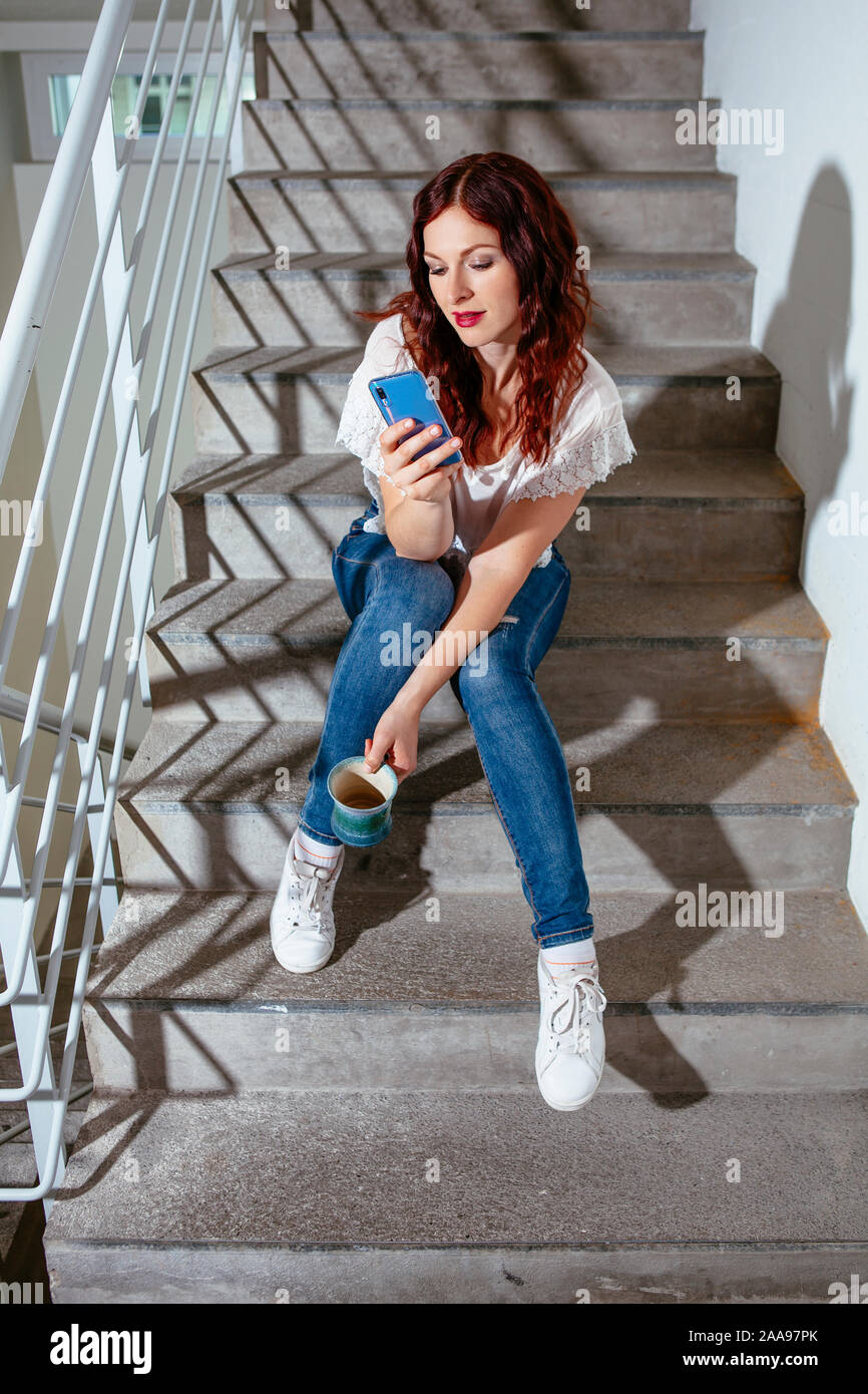 Giovane donna con i capelli rossi seduta sulle scale e parlando sul suo smartphone e di bere il caffè. Foto Stock