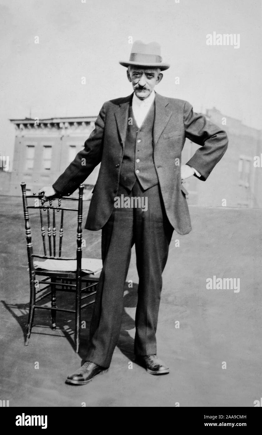 Un stern cerca uomo di mezza età colpisce insolente posa mentre stringendo un periodo sedia in New York City rooftop, ca. 1930. Foto Stock