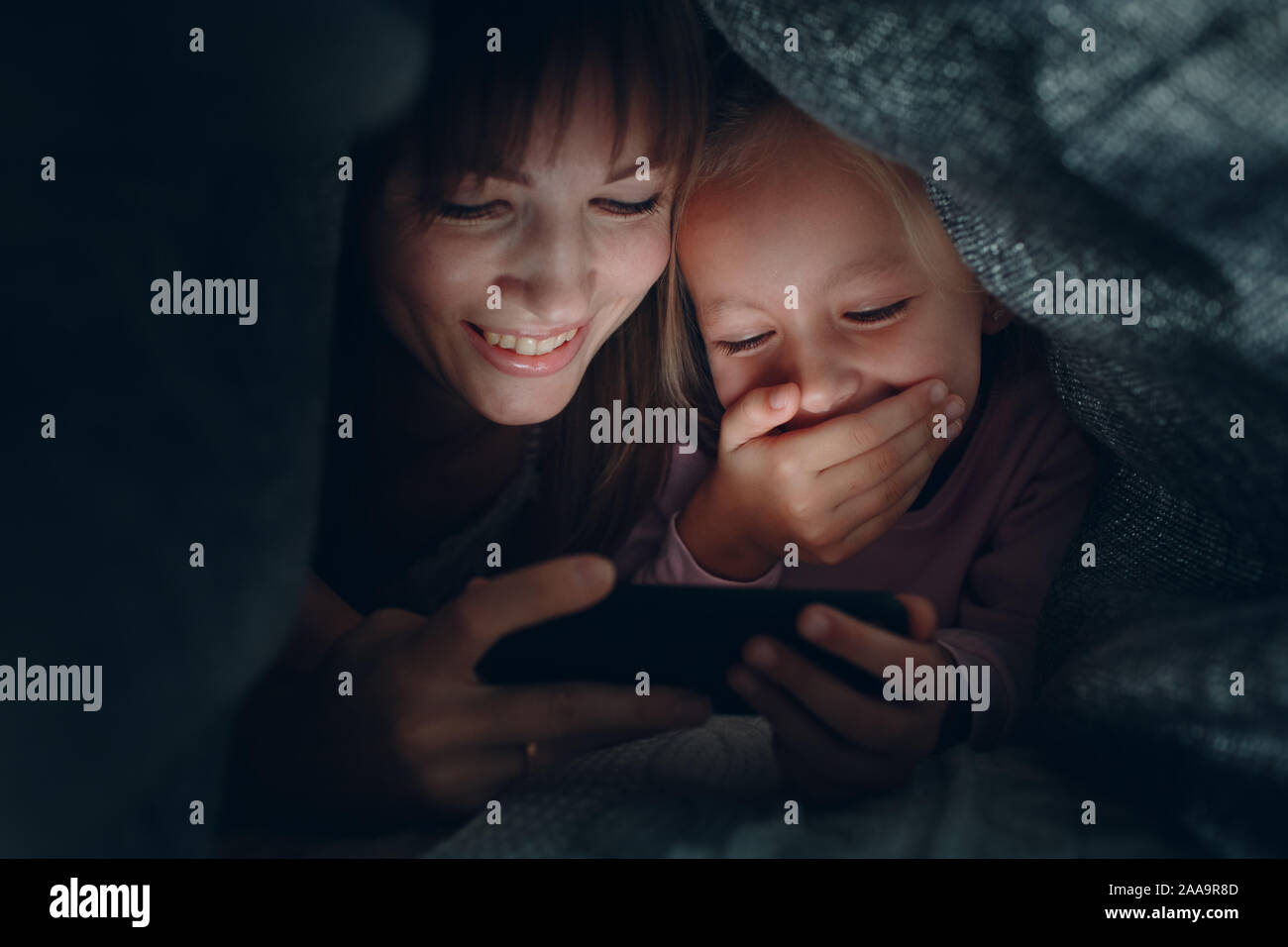 Madre di una bambina guarda il contenuto sullo smartphone nel buio sotto la coperta copre. Foto Stock