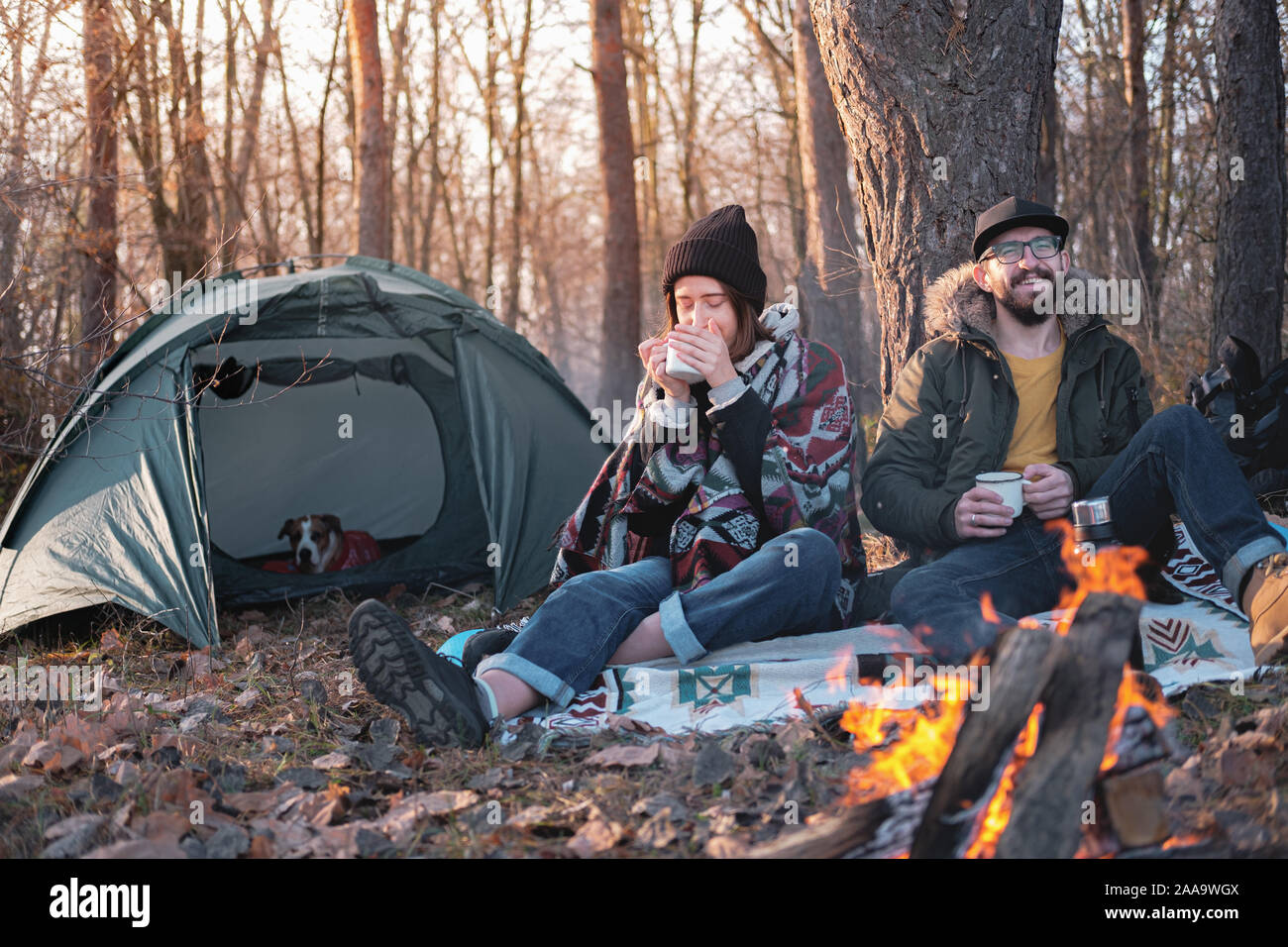 Due persone che godono di bevande calde per il falò in campeggio. Andare a fare un escursione in stagione fredda: uomo e donna sedere accanto al fuoco con una tenda in bac Foto Stock
