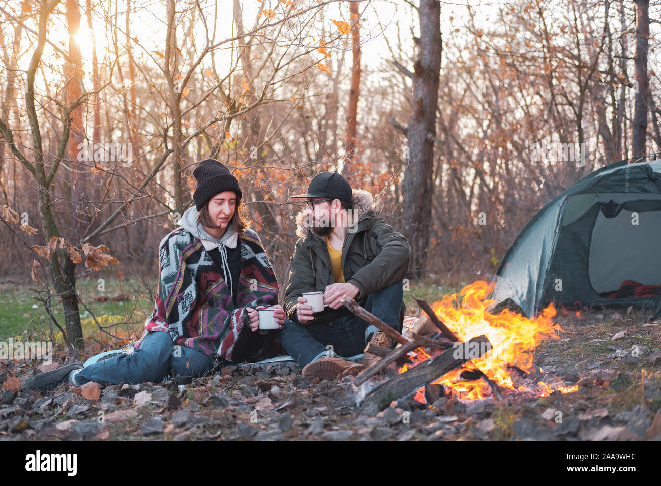 Due persone che godono di bevande calde per il falò in campeggio. Andando su un trekking vacanza in stagione fredda: uomo e donna sedere accanto al fuoco con un te Foto Stock