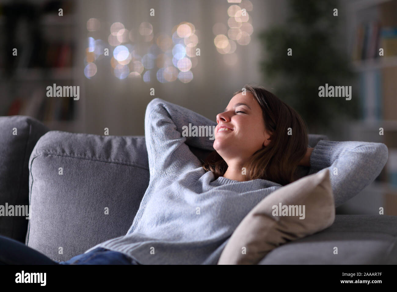 Donna felice rilassante con le braccia sulla testa seduto su un divano nella notte a casa Foto Stock