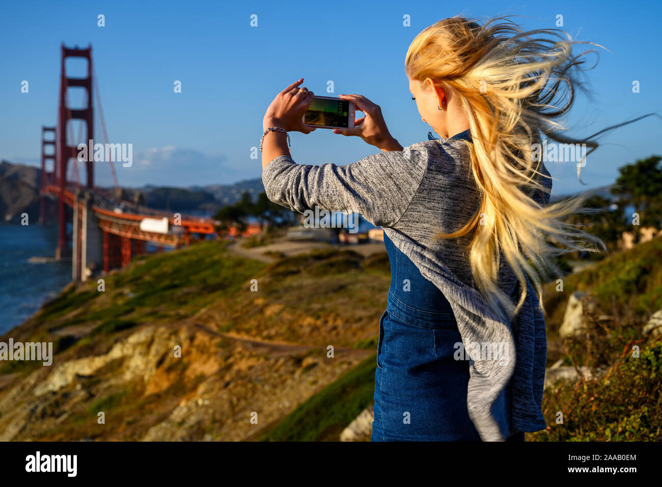 Attraente ragazza bionda di scattare le foto del Golden Gate Bridge e i suoi lunghi capelli biondi è volare nel vento forte a San Francisco, California, Stati Uniti d'America Foto Stock