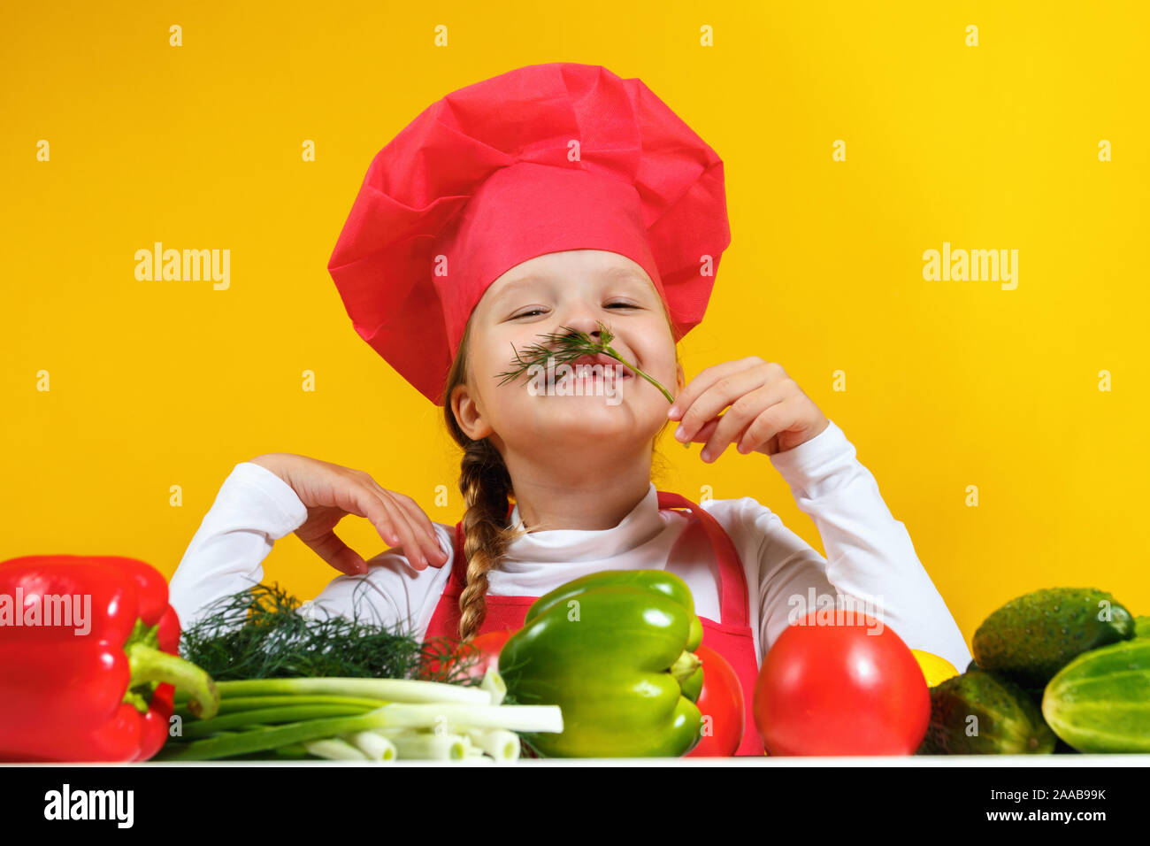 Bellissima bambina chef su sfondo giallo. Un bambino prepara una insalata di verdure, ingannare intorno e facendo un baffi di aneto. Foto Stock