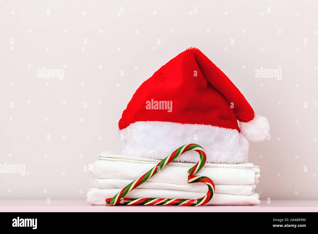 Primo piano di una pila di pulire biancheria da letto bianca, Santa hat e Natale caramelle, sullo sfondo di una parete di luce. Copia dello spazio. Foto Stock