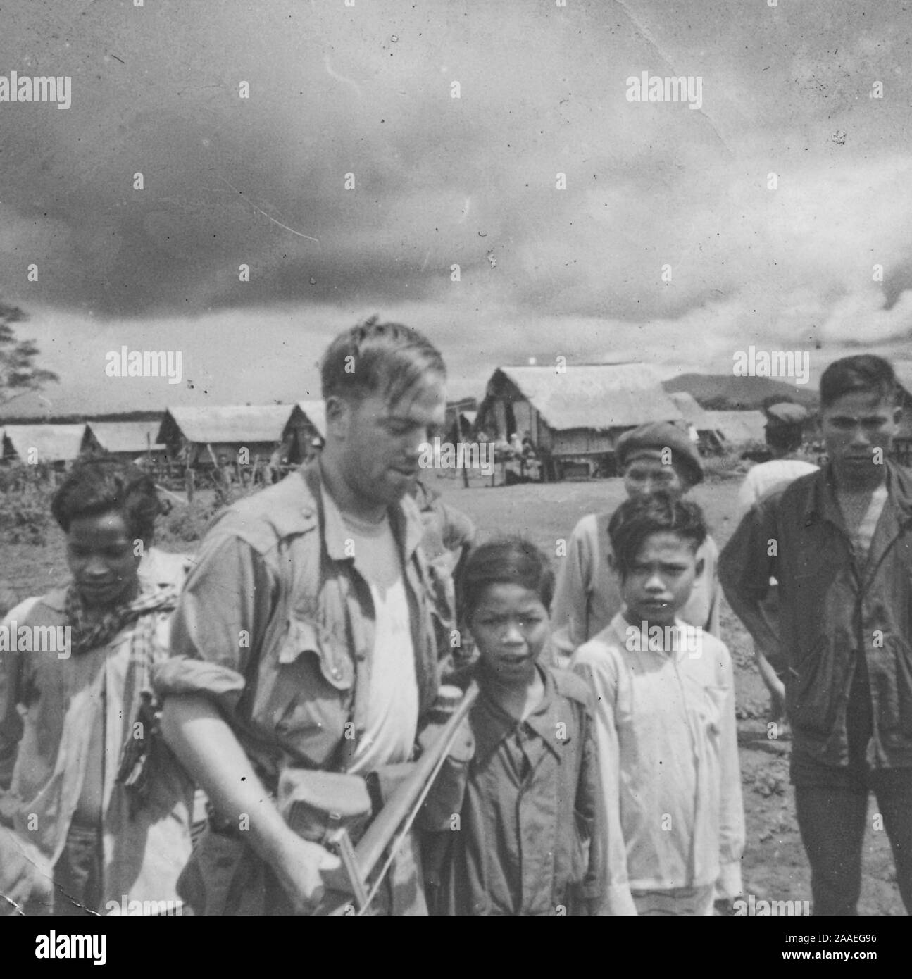 Close-up di un veterano americano, tenendo un lungo attrezzo fatti a mano e di stare in mezzo a molti ragazzi vietnamiti e gli uomini, in un aperto campo agricolo con tetto in paglia con stilt-case in background, Vietnam, 1966. () Foto Stock
