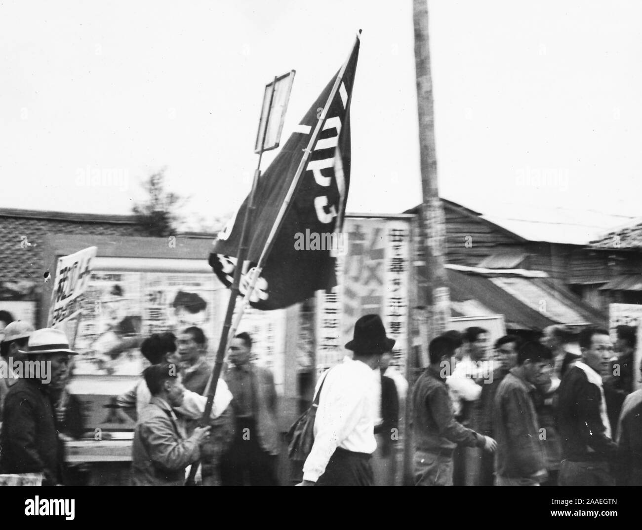 Vista di profilo di uomini, alcune recanti segni e una grande bandiera, marciando attraverso una zona urbana in un giorno nuvoloso durante la partecipazione della manodopera europea proteste, Prefettura di Fukuoka, Giappone, 1950. () Foto Stock