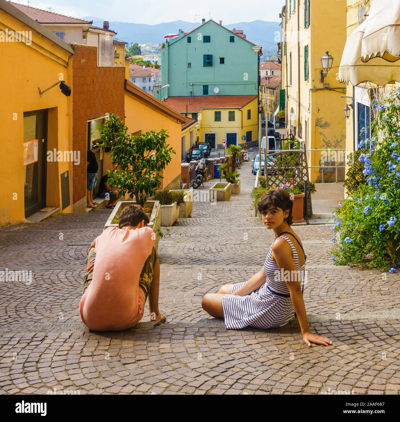 Un ragazzo e una ragazza abbronzata seduti in strada, riposati nella vecchia città italiana di Imperia, con colorate case pastello sullo sfondo Foto Stock