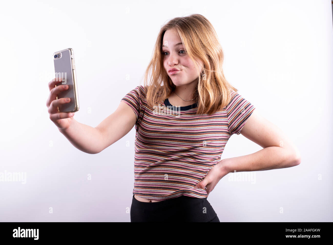 Caucasian ragazza adolescente che indossa un rigato orizzontale T-shirt con un espressione divertente tenendo un selfie con uno smartphone contro uno sfondo bianco Foto Stock