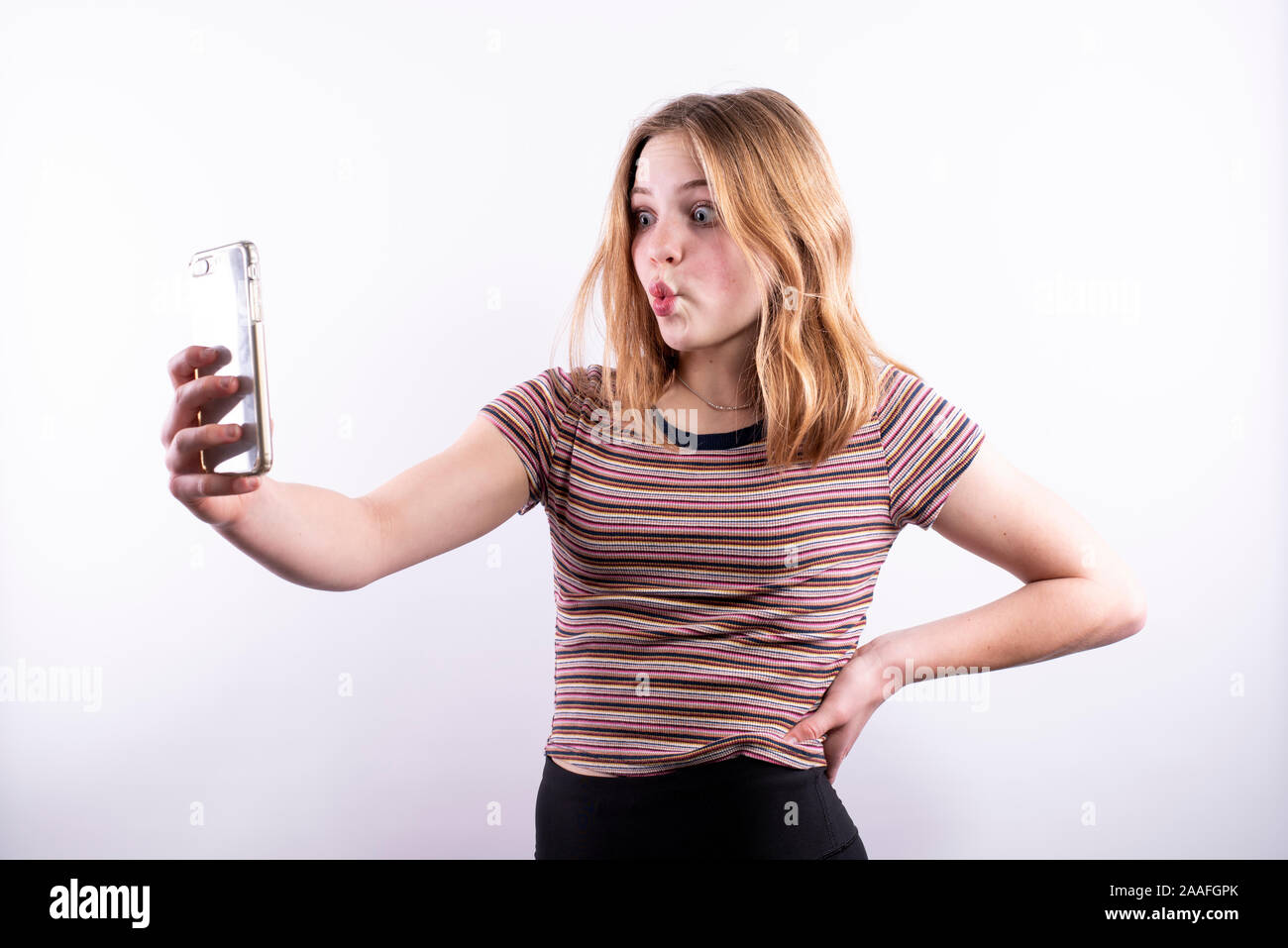 Caucasian ragazza adolescente che indossa un rigato orizzontale T-shirt con un bacio espressione e occhi spalancati mentre prendendo un selfie con uno smartphone Foto Stock