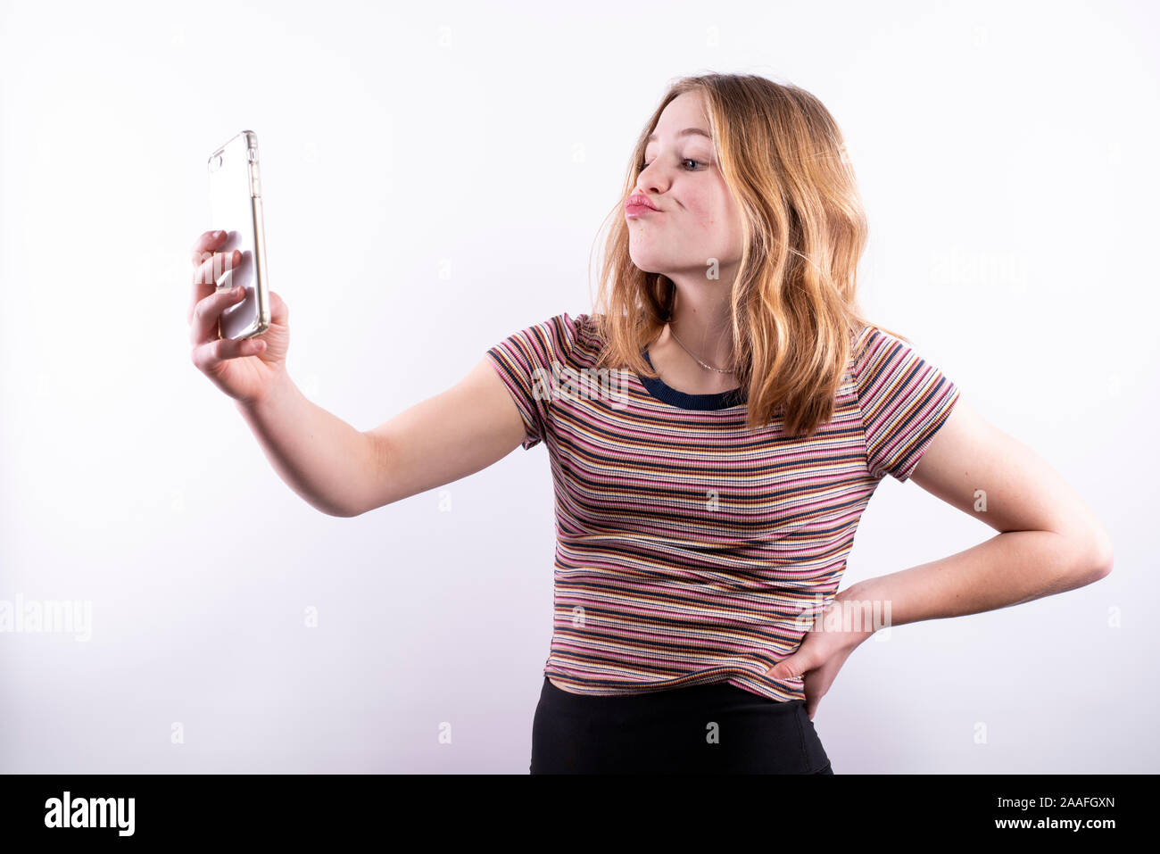 Caucasian ragazza adolescente che indossa un rigato orizzontale T-shirt con un espressione divertente tenendo un selfie con uno smartphone contro uno sfondo bianco Foto Stock