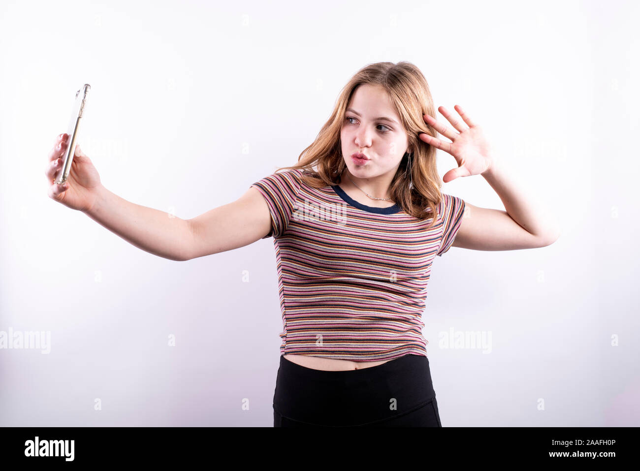 Caucasian ragazza adolescente che indossa un rigato orizzontale T-shirt prendendo un drammatico selfie con uno smartphone contro uno sfondo bianco Foto Stock