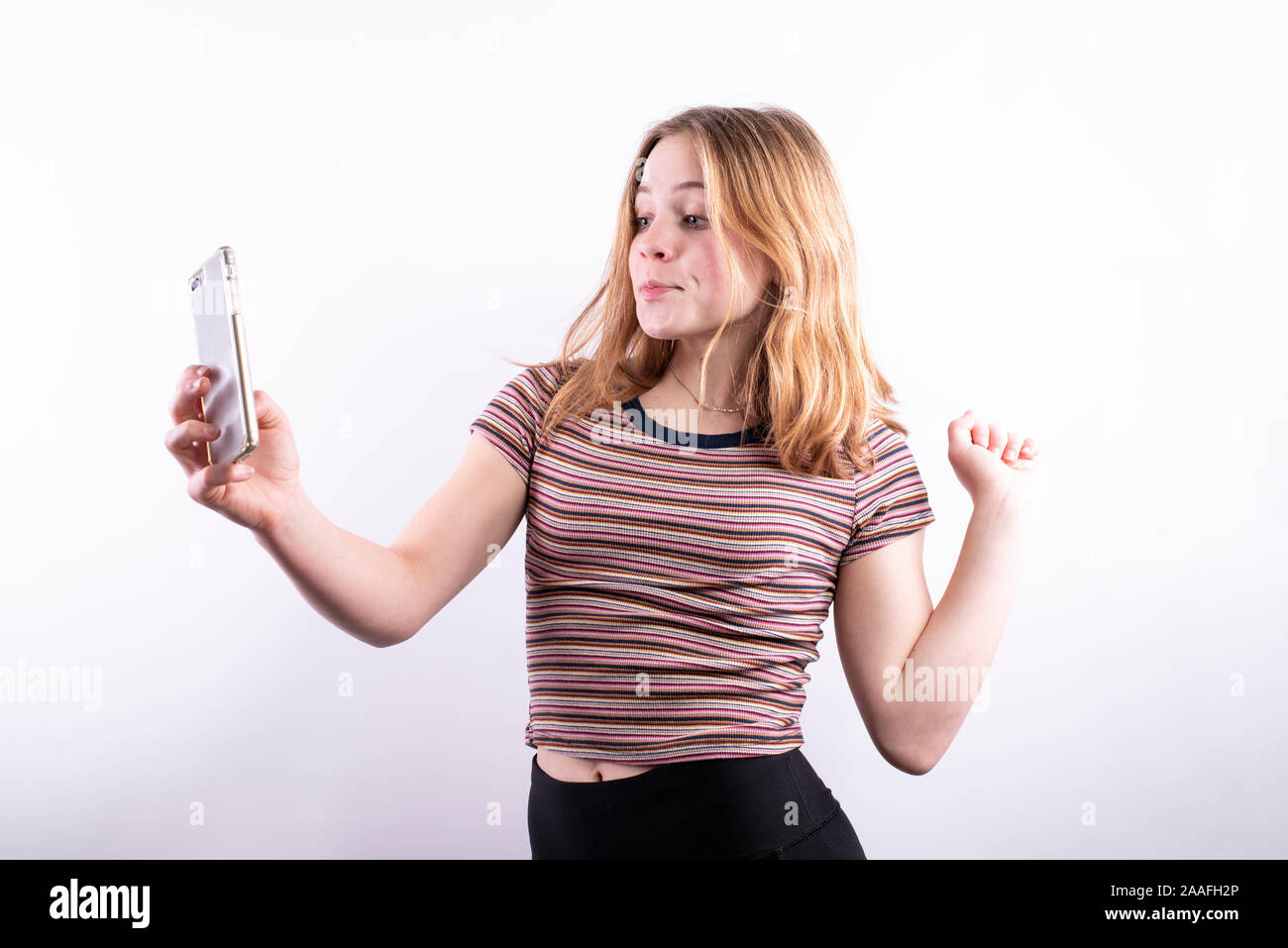 Caucasian ragazza adolescente che indossa un colorato a strisce orizzontali T-shirt con un espressione divertente e pursed lips tenendo un selfie con uno smartphone Foto Stock