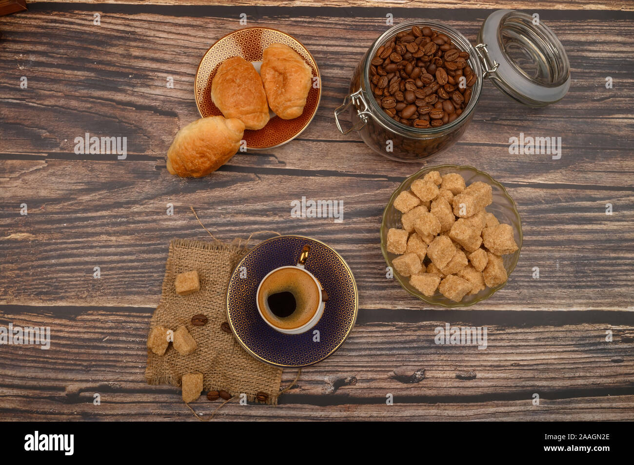 Una tazza di caffè, pezzi di zucchero di canna in una zuccheriera, i chicchi di caffè in un vasetto di vetro e croissant su uno sfondo di legno Foto Stock